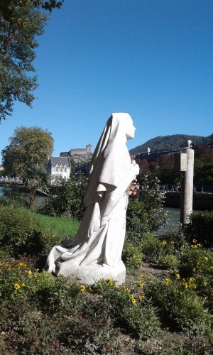 statue blanche de Bernadette Soubirou à genoux priant en direction de la grotte de Lourde de l'autre coté du torrent